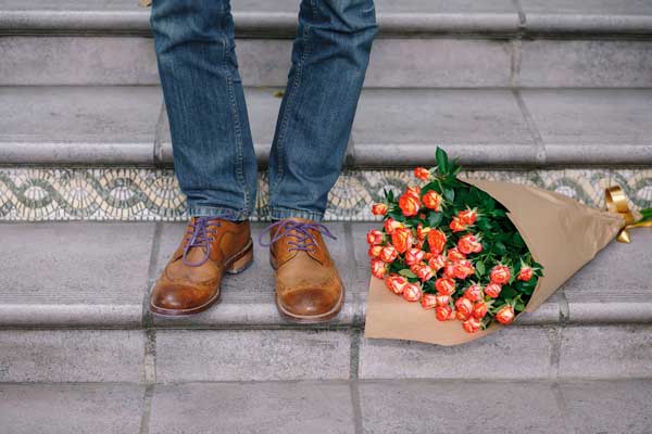 The bottom of a pair of legs in jeans and brown shoes, next to a bunch of flowers